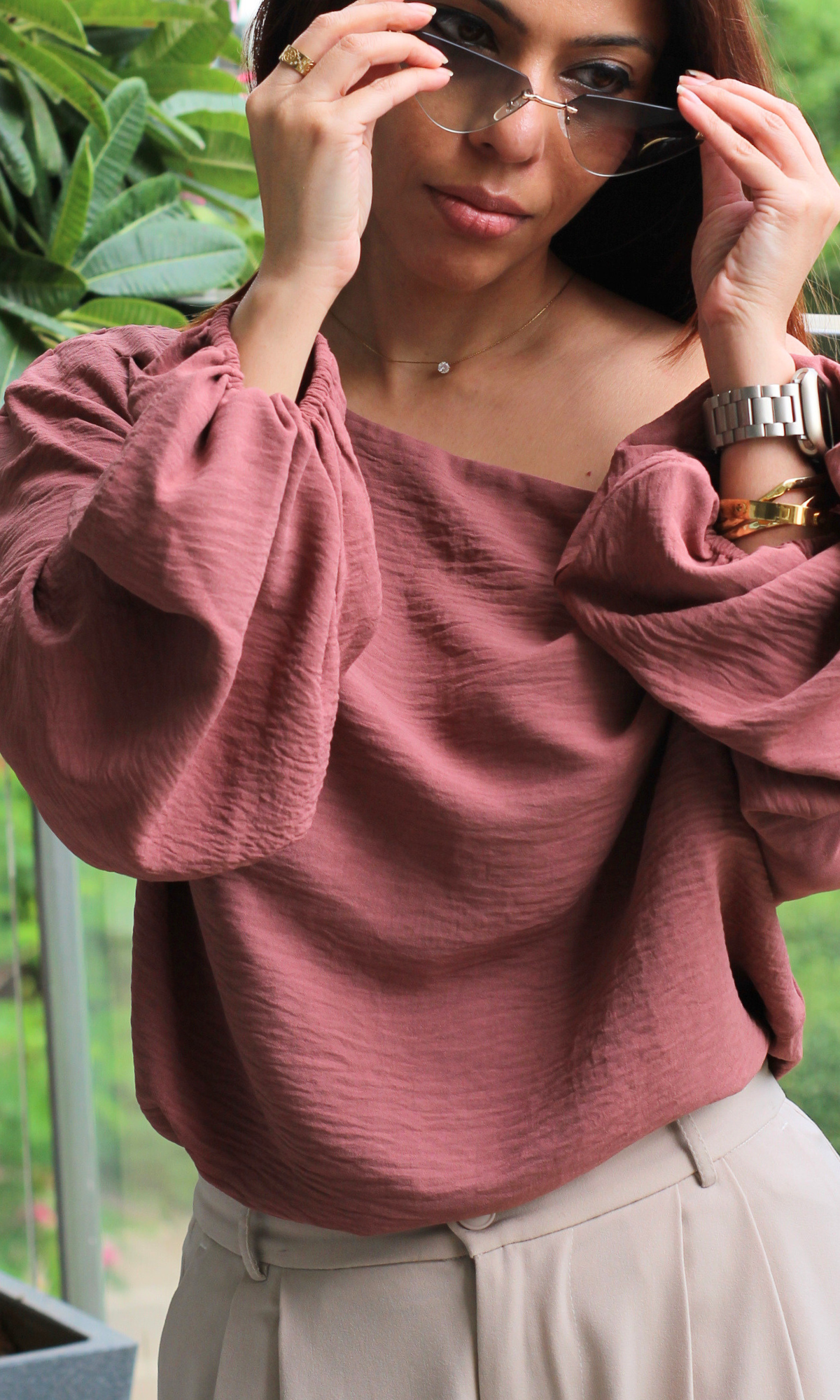 Woman in a mauve off-shoulder blouse with relaxed sleeves and cream pleated trousers, styled with sunglasses and a brown shoulder bag, posing on a balcony.
