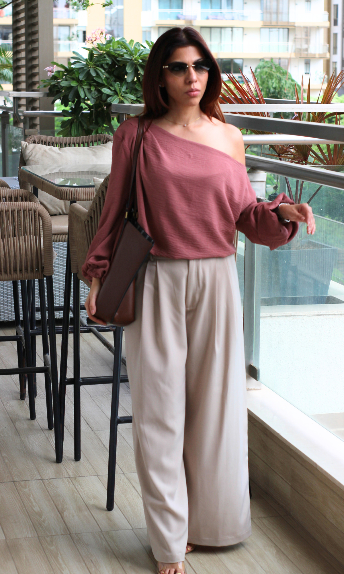 Chic mauve off-shoulder blouse paired with cream trousers and a brown bag, styled with minimal jewelry and sunglasses on a balcony.