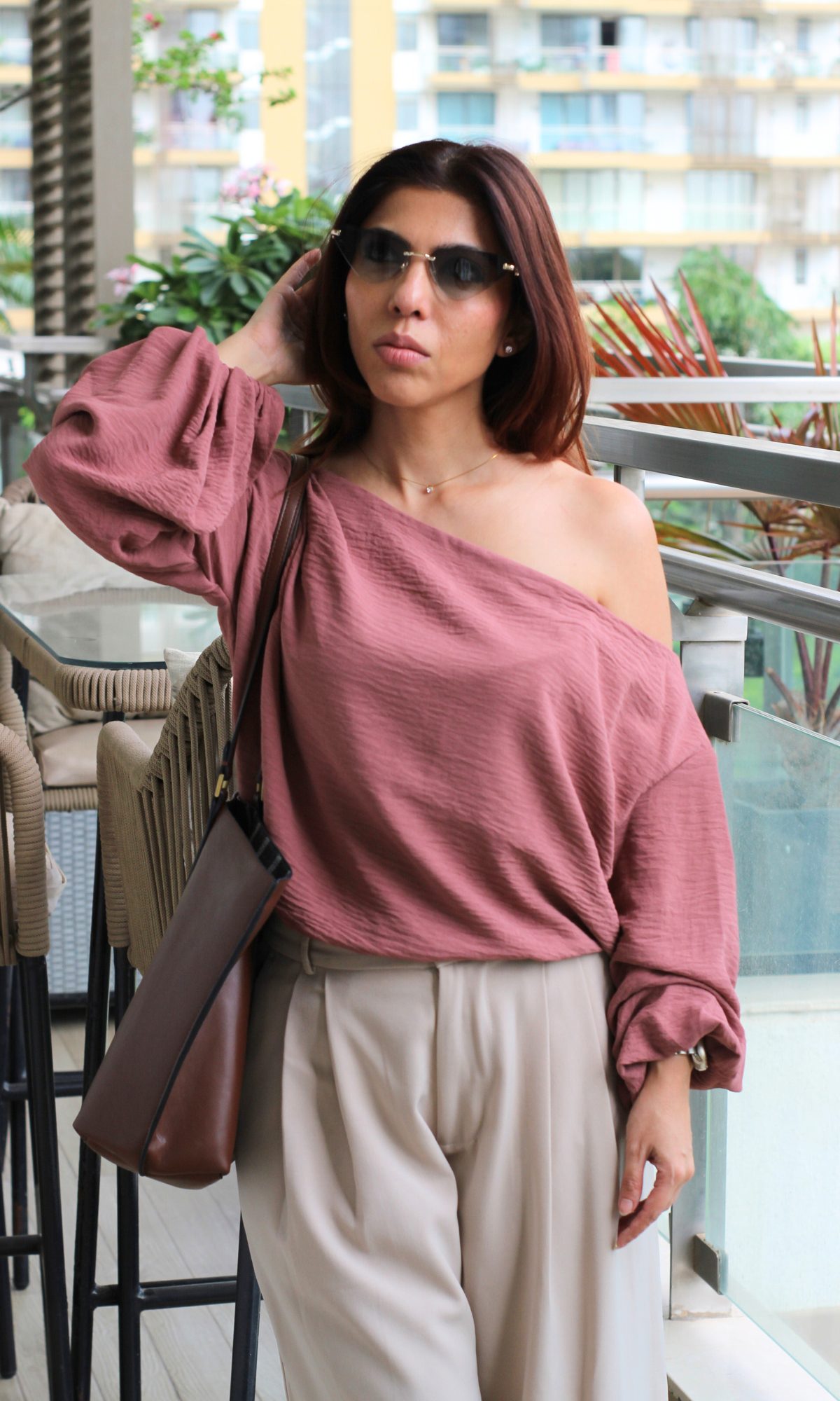Mauve off-shoulder blouse with cream pleated pants and brown shoulder bag, styled with sunglasses.