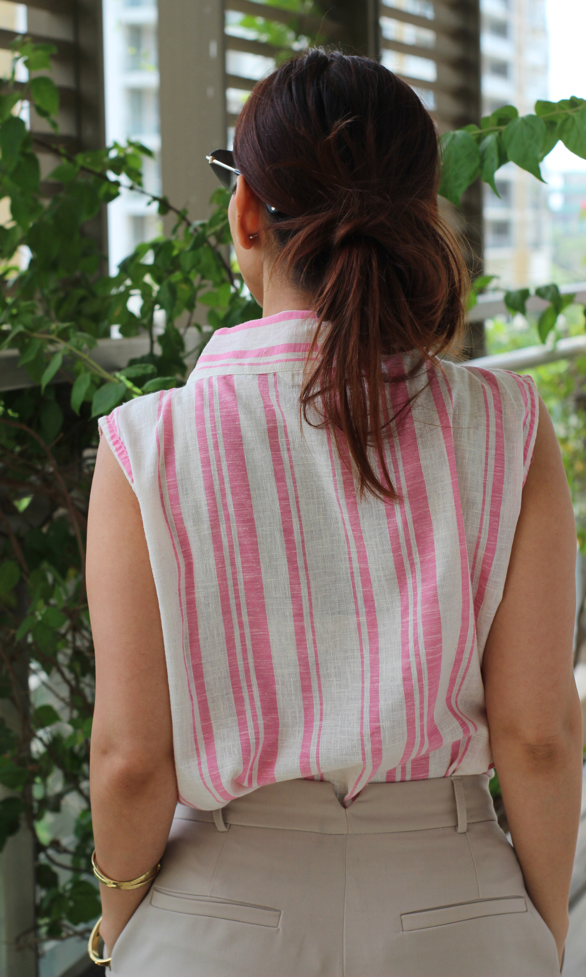 Back view of a woman wearing a sleeveless pink and white striped cotton top paired with beige high-waisted trousers, standing outdoors.