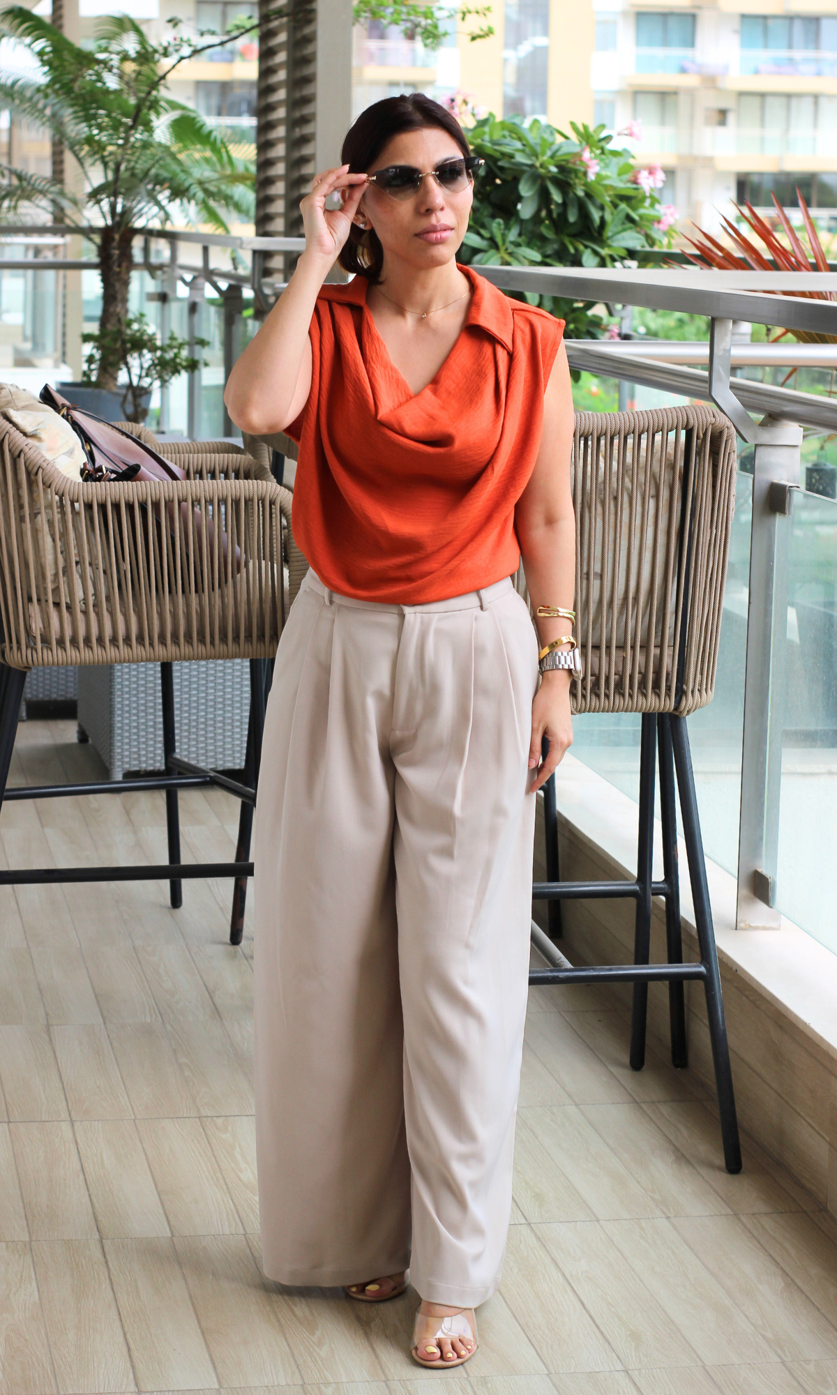 Woman wearing a sleeveless rust-orange draped cowl-neck top paired with high-waisted beige pleated trousers.