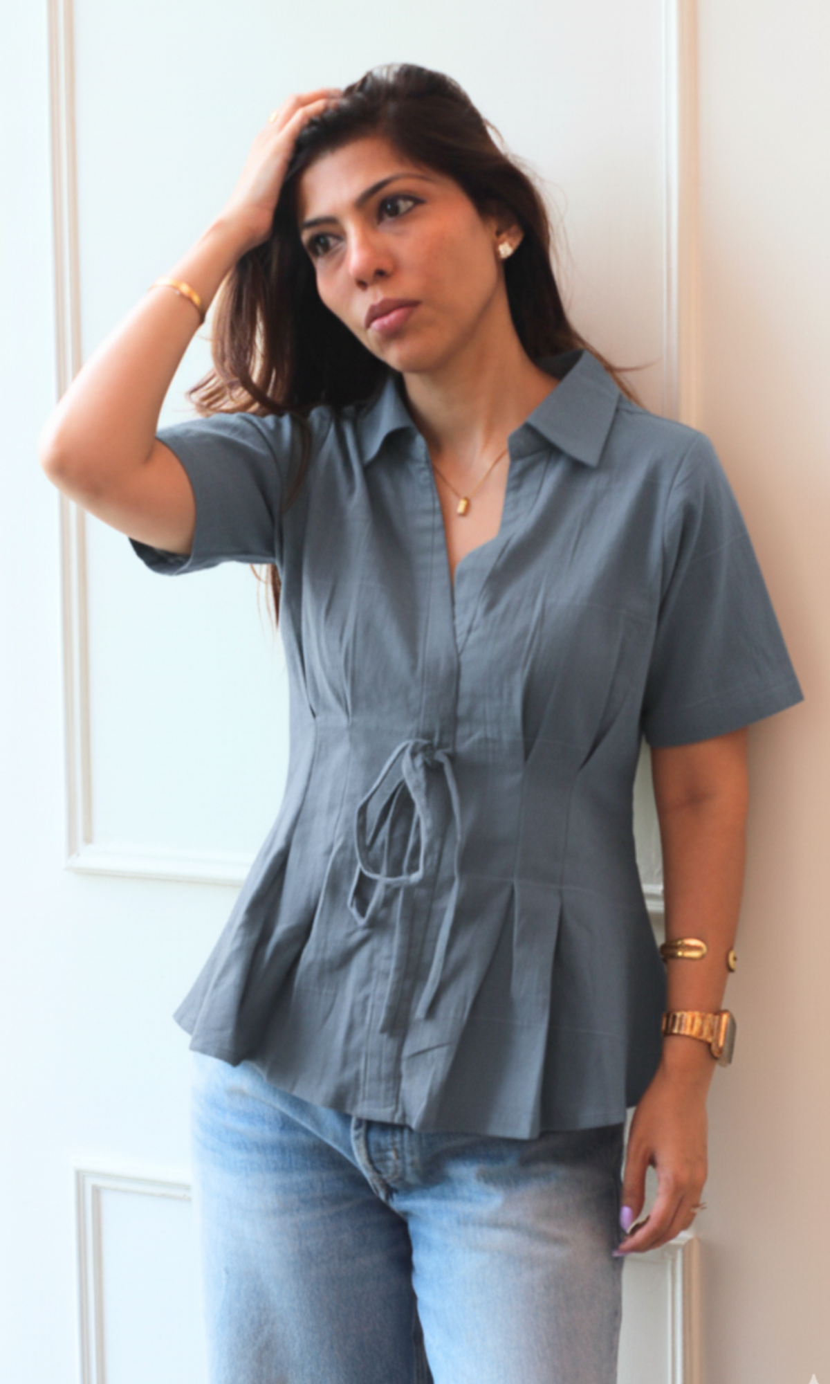 Stylish woman in a short-sleeve grey tie-front blouse and light denim jeans, accessorized with gold jewelry and sunglasses, posing confidently in an urban setting.