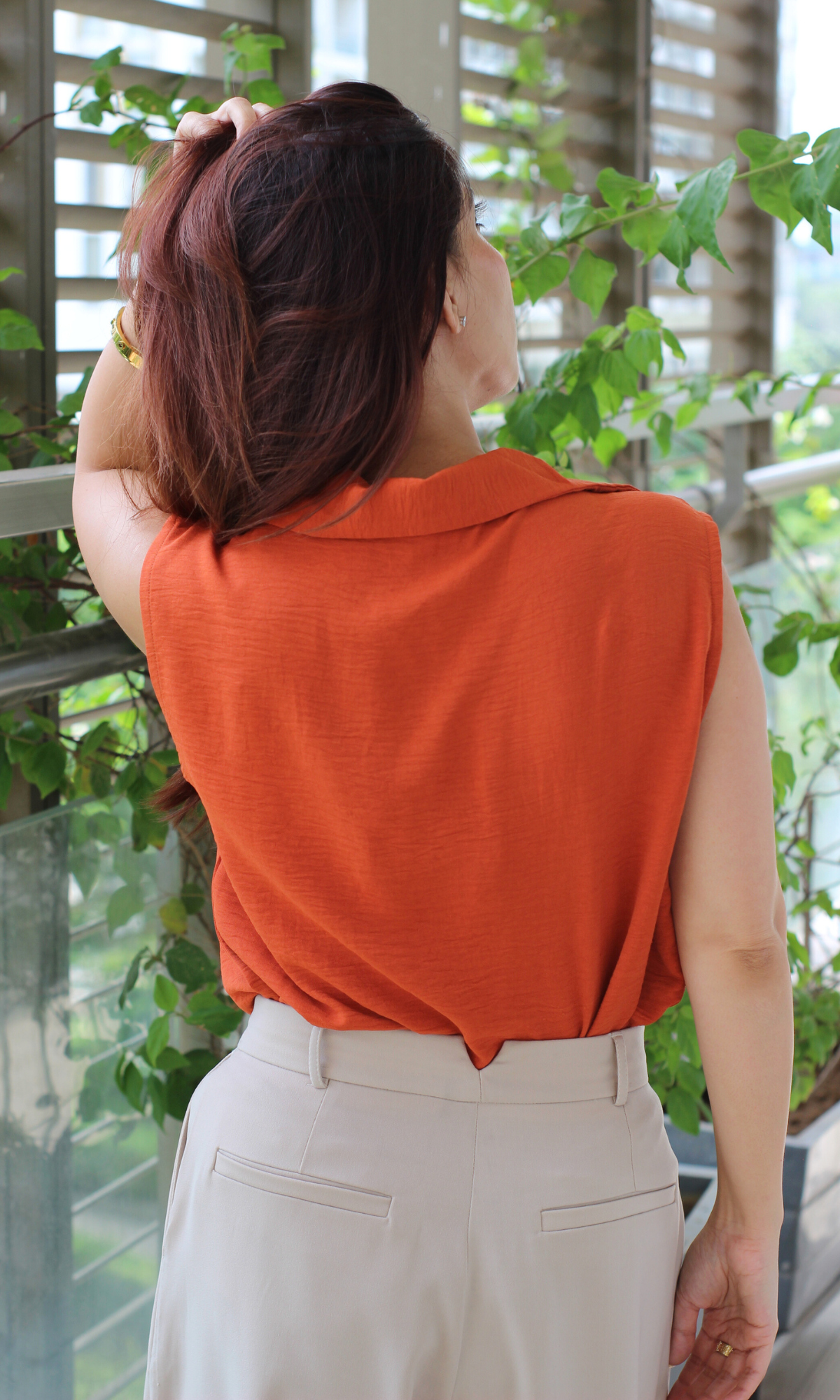 Back close-up of a sleeveless rust-orange draped cowl-neck top showing the soft fabric and relaxed fit, paired with beige pleated trousers.