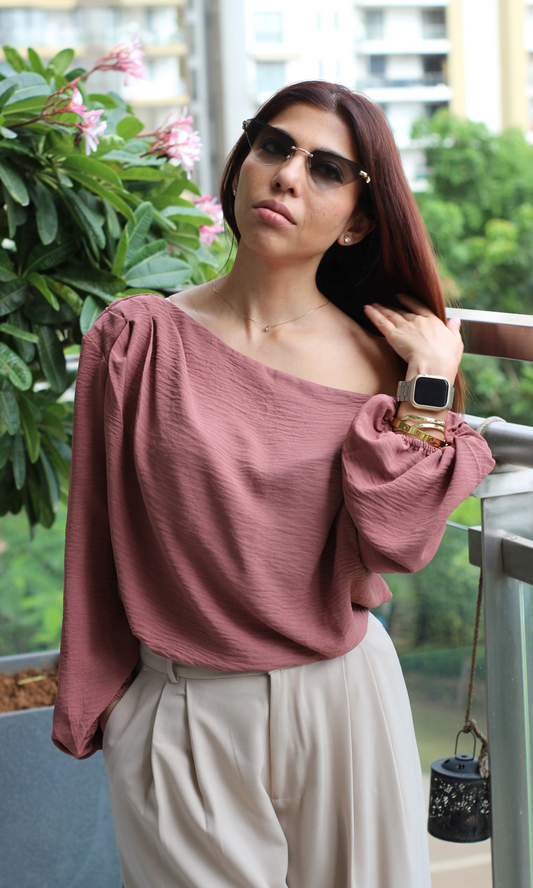 Modern casual outfit with a Burnt Umber off-shoulder textured top, cream pleated trousers, sunglasses, and a brown crossbody bag, styled for a balcony look.