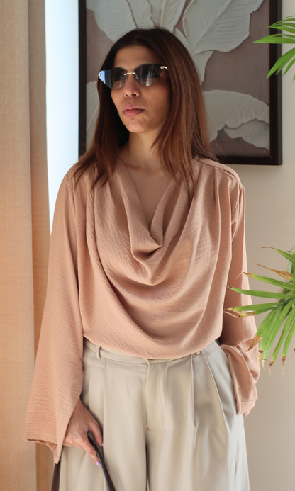 Model wearing a dusky rose-colored top featuring a draped cowl neckline and full sleeves with slit details, styled in a cozy indoor setting.