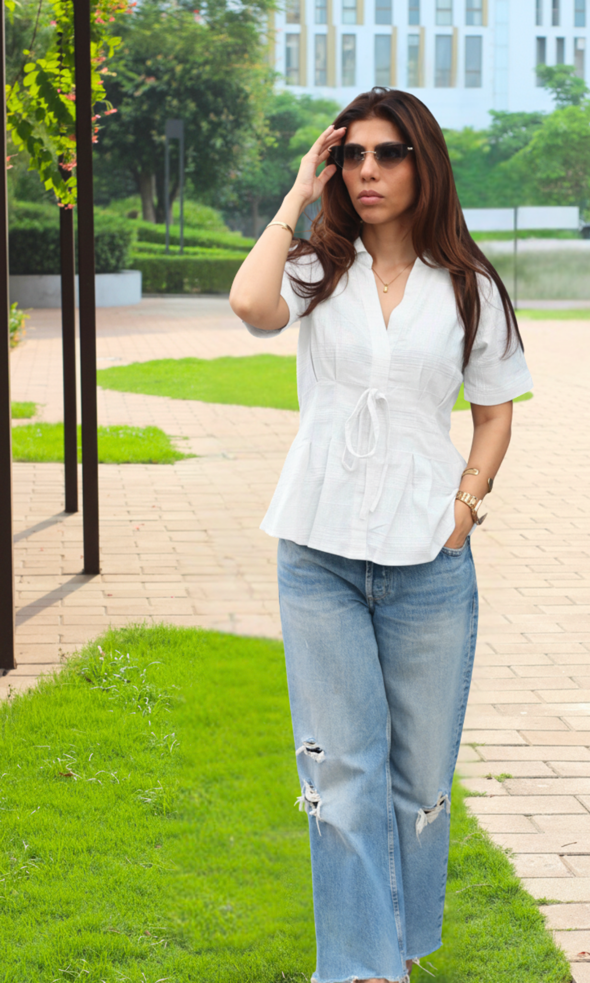 Elegant woman in a white V-neck tie-front top and casual blue jeans, styled with gold accessories, posing indoors against a clean white backdrop.